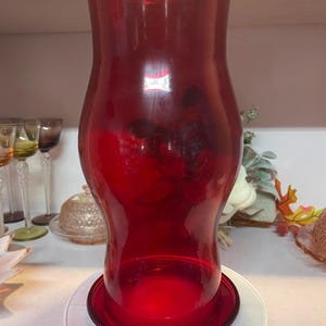 May include: A tall, ruby red glass vase with a flared base and a wide, curved body. The vase sits on a clear, round base. The glass is transparent, allowing light to pass through. The vase is likely used for decorative purposes.