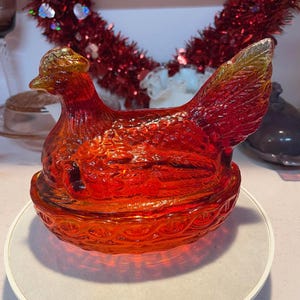 May include: A vibrant, red-orange glass hen-shaped trinket box. The lid and base are textured with a basket weave design. The hen's head and tail feathers have a golden hue. A decorative piece for the home.