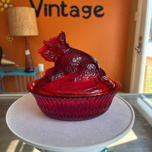 May include: A ruby red glass candy dish featuring a sculpted dog figurine as the lid handle. The bowl has a ribbed, vertical pattern along the exterior sides, while the lid is shaped like a seated terrier. The glass is translucent and deep red.