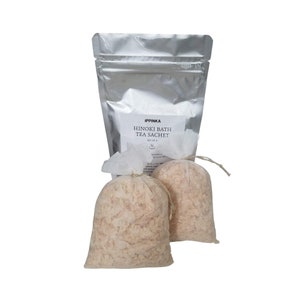 May include: Two white muslin bags filled with a light brown bath tea blend. The bags are tied with twine and are sitting on a silver foil bag with the text "IPPINKA HINOKI BATH TEA SACHET SET OF 2".