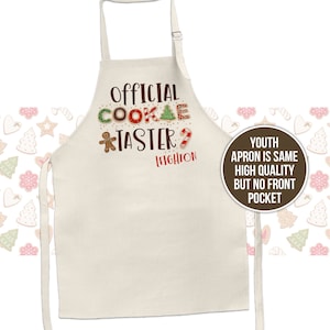 May include: A beige youth apron with the text "Official Cookie Taster" in red, green, and brown. The text is decorated with gingerbread cookie images. The name "Leighton" is written in brown below the text. The apron has adjustable straps and a brown circle with the text "Youth Apron is same high quality but no front pocket" in white.