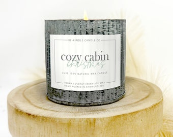 May include: A gray metal candle jar with a white label that reads "RE-KINDLE CANDLE CO. cozy cabin Christmas LUXE 100% NATURAL WAX CANDLE VEGAN COCONUT CREAM SOY WAX HAND POURED IN KIRKWOOD, MO". The candle is lit and sitting on a wooden stand.