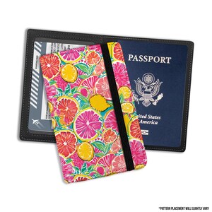 May include: A passport holder featuring a vibrant pattern of pink, yellow and green citrus fruits. The holder has a black elastic band to secure the passport. The text "PASSPORT" is visible on the blue passport inside the holder.