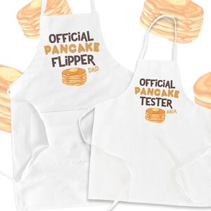 May include: Two white aprons with brown text and images of pancakes. The text on the first apron reads "Official Pancake Flipper Dad". The text on the second apron reads "Official Pancake Tester Aria".