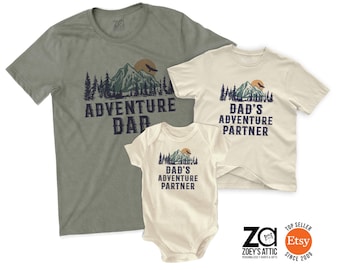 May include: Three matching t-shirts and a onesie with mountain and sun graphics. The adult shirts say "Adventure Dad" and "Dad's Adventure Partner." The onesie also says "Dad's Adventure Partner."