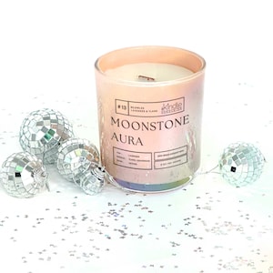 May include: A pink and iridescent glass candle jar with a wooden wick. The candle is labeled "#13 BEJEWLED LAVENDER & YLANG" and "MOONSTONE AURA". The candle is surrounded by silver disco ball ornaments on a white background.