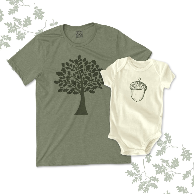 May include: A matching set of green and white t-shirts featuring a tree and an acorn design. The adult t-shirt has a green tree with a brown trunk and the baby t-shirt has a white acorn with a brown cap.