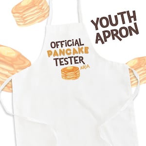 May include: White youth apron with the text "OFFICIAL PANCAKE TESTER" in brown letters. The text "YOUTH APRON" is in brown letters at the top right corner. The name "ARIA" is written in smaller brown letters below the text "OFFICIAL PANCAKE TESTER". There is a cartoon image of a stack of pancakes with butter on top.