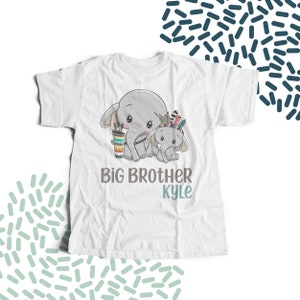 May include: White t-shirt featuring a graphic of two elephants. The larger elephant is wearing a colourful headdress and holding a cup. The smaller elephant is standing next to the larger elephant. The text "Big Brother Kyle" is printed below the elephants.
