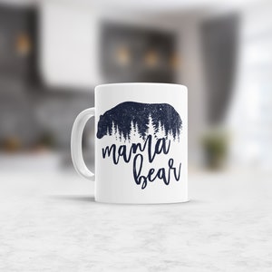 May include: White ceramic mug with a black silhouette of a bear with trees inside the bear. The text "mama bear" is written below the bear.