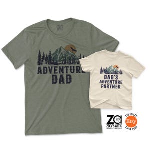 May include: Two short-sleeved t-shirts. One is olive green with the text "ADVENTURE DAD" and a mountain graphic. The other is off-white with "DAD'S ADVENTURE PARTNER" and a similar mountain design. Both shirts have a round neckline.