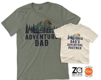 May include: Two short-sleeved t-shirts. One is olive green with the text "ADVENTURE DAD" and a mountain graphic. The other is off-white with "DAD'S ADVENTURE PARTNER" and a similar mountain design. Both shirts have a round neckline.