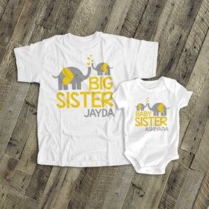 May include: Two white t-shirts featuring grey and yellow elephant designs. The larger shirt reads "Big Sister Jayda" and the smaller shirt reads "Baby Sister Ashiyana".