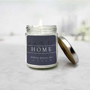 May include: A clear glass jar candle with a silver lid. The candle has a white label with the text "Cozy Home" and "webster groves, mo 63119" printed in black.
