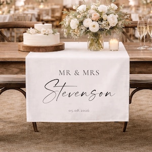 Puede incluir: Una mesa de recepción de bodas con un camino de mesa blanco impreso con "MR & MRS Stevenson" y la fecha 05.08.2026. La mesa está decorada con un pastel, flores, velas y copas de champán. El fondo muestra otras mesas y decoraciones.