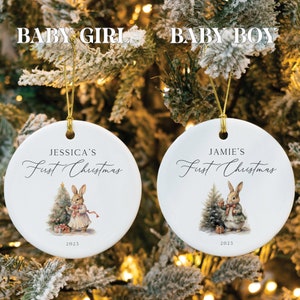 以下が含まれることがあります： ウサギのイラストと「First Christmas」の文字が書かれた白いセラミック製のクリスマスオーナメントが2つ。左側のオーナメントには「Jessica's」、右側のオーナメントには「Jamie's」と書いてあります。両方のオーナメントには2023年が印刷されています。