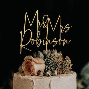Op de afbeelding: Een tweelaagse taart met wit glazuur, versierd met verse rozen en groen, bekroond met een goudkleurige taarttopper in cursief schrift met de tekst Mr & Mrs Robinson tegen een effen zwarte achtergrond.