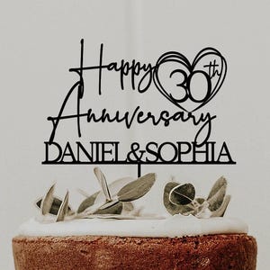 May include: A tiered cake with white frosting and foliage, topped with a black cake topper. The topper says "Happy Anniversary" with "30th" in a heart, and the names "DANIEL & SOPHIA".