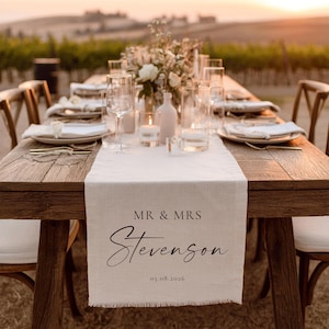 May include: A wooden table set for a wedding reception. A cream table runner with the text "MR & MRS Stevenson" and the date 05.08.2026 is centered. The table is set with plates, glasses, and candles, with a floral centrepiece.