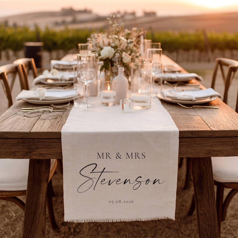 May include: A wooden table set for a wedding reception. A cream table runner with the text "MR & MRS Stevenson" and the date 05.08.2026 is centered. The table is set with plates, glasses, and candles, with a floral centrepiece.