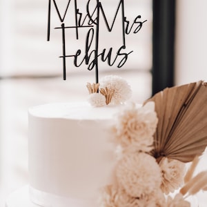 May include: A two-tiered white wedding cake with a black acrylic topper that reads "Mr & Mrs Febus". The cake is decorated with white and beige dried flowers.