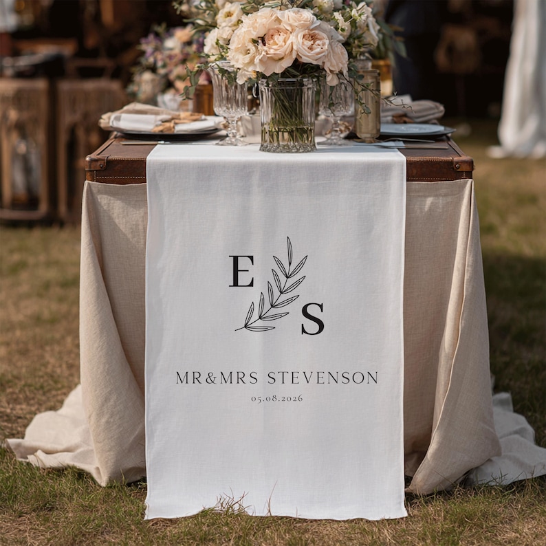 May include: A white linen table runner featuring the initials "E" and "S" in black, accompanied by a botanical illustration. The text "MR & MRS STEVENSON" and the date "05.08.2026" are printed on the runner. A floral display is visible in the background.