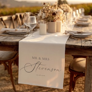 May include: A wooden table set for a wedding reception. A cream table runner displays the text "MR & MRS Stevenson" and the date 05.08.2026. The table is set with plates, glasses, candles, and floral arrangements.