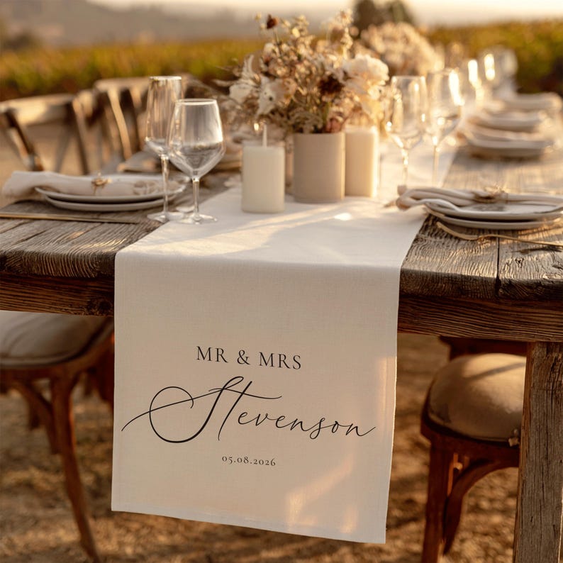 May include: A wooden table set for a wedding reception. A cream table runner displays the text "MR & MRS Stevenson" and the date 05.08.2026. The table is set with plates, glasses, candles, and floral arrangements.