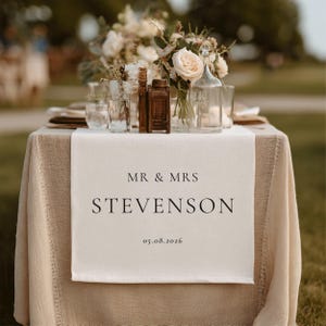 May include: A wedding table setting with a beige linen tablecloth and a white runner. The runner displays the text "MR & MRS STEVENSON" and the date "05.08.2026". The table is decorated with floral arrangements and glassware.