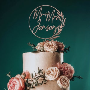 May include: A two-tiered wedding cake decorated with blush pink roses and greenery. A wooden cake topper with the text "Mr & Mrs. Johnson" in a script font is on top of the cake.