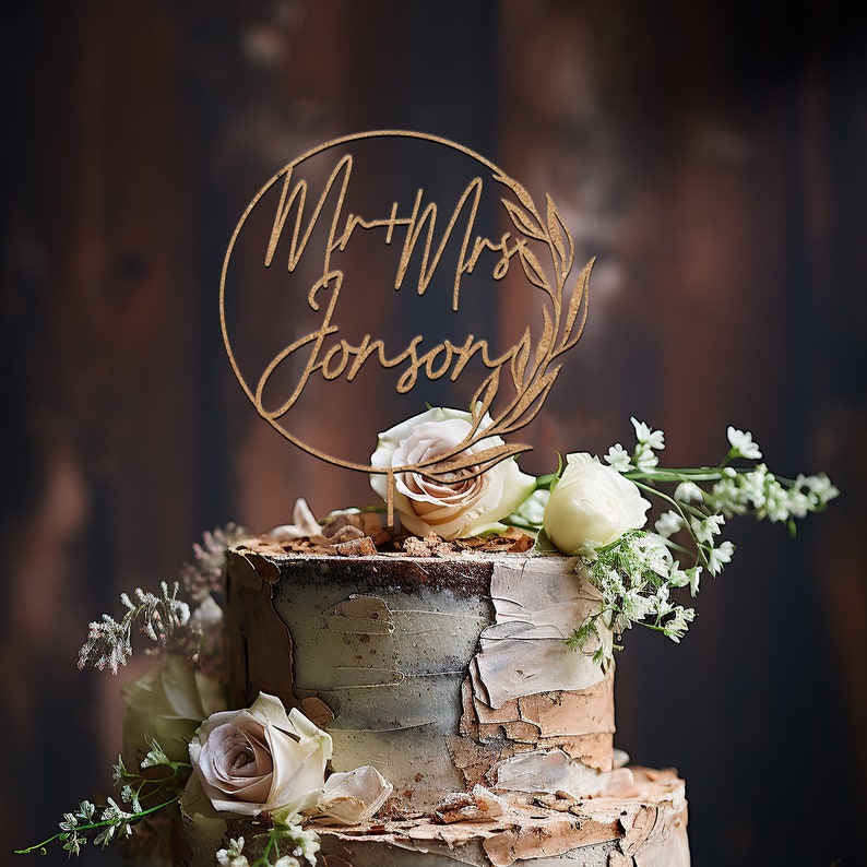 May include: A wooden cake topper with the text "Mr. & Mrs. Johnson" in a circle with leaves. The topper is on a two-tiered cake decorated with white and brown frosting and flowers.