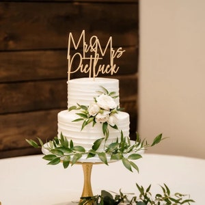 May include: A two-tiered white wedding cake decorated with white flowers and greenery. The cake topper says "Mr. & Mrs. Prettluck".