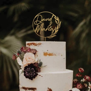 May include: Gold wedding cake topper with the names "David & Maria" in a cursive font, surrounded by a wreath of leaves. The topper is on a white cake with burgundy and pink flowers.