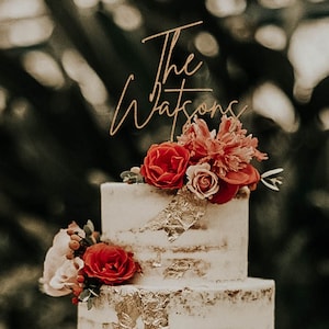 May include: A two-tiered wedding cake decorated with red and pink flowers and gold leaf. The cake topper says "The Watsons" in gold script.