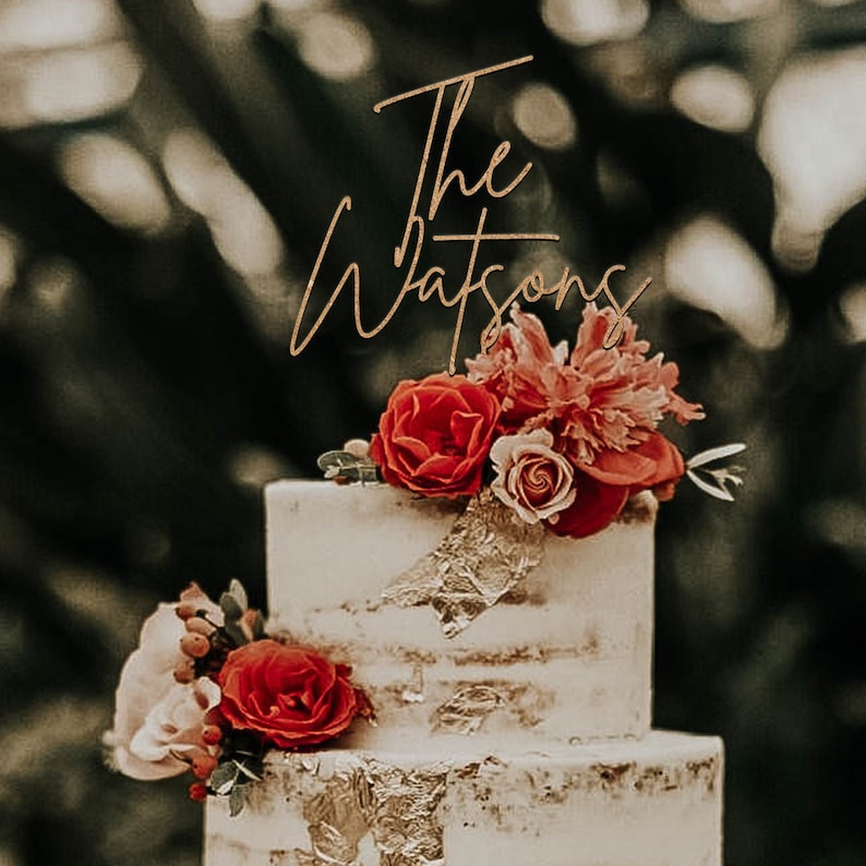 May include: A two-tiered wedding cake decorated with red and pink flowers and gold leaf. The cake topper says "The Watsons" in gold script.