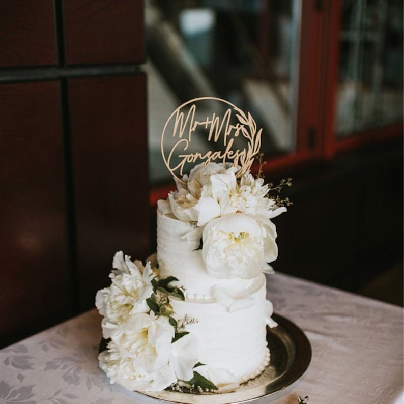 May include: A two-tiered white wedding cake decorated with white peonies and greenery. The cake topper is a wooden circle with the text "Mr. & Mrs. Gonzales" in cursive.