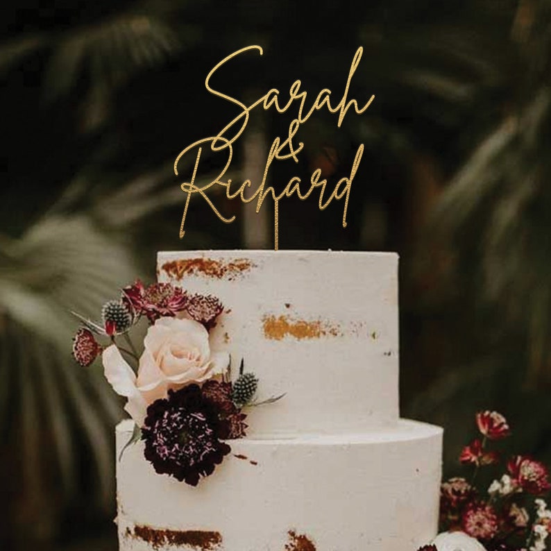 May include: A two-tiered white wedding cake with gold accents and burgundy flowers. A gold glitter topper reads "Sarah & Richard".
