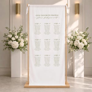 Pu&ograve; includere: Un cartello in tessuto bianco per il tableau de mariage, sospeso su un supporto in legno chiaro. Il cartello presenta il testo "OUR FAVORITE PEOPLE" e "please find your seat" in lettere dorate, seguito da nove tavoli con elenchi di nomi in grigio scuro.