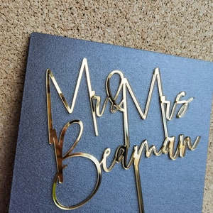 May include: Gold Mr & Mrs Bedman cake topper with a cursive font. The topper is on a dark gray background.