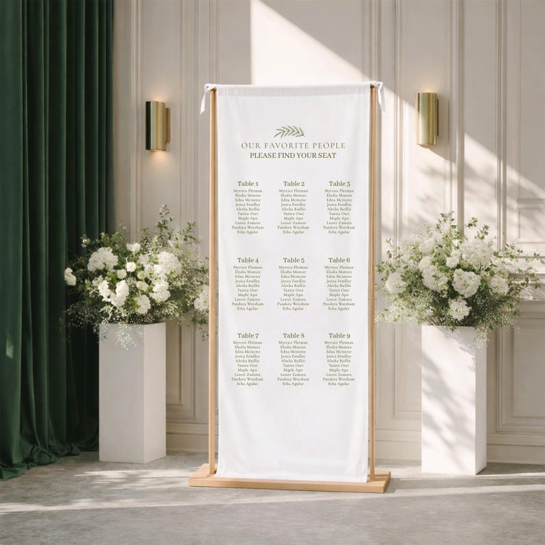 Pu&ograve; includere: Un alto cartello rettangolare per il tableau de mariage in tessuto bianco, sospeso a una cornice in legno chiaro. Il cartello riporta il testo "OUR FAVORITE PEOPLE" e "PLEASE FIND YOUR SEAT" sopra un elenco di nove tavoli con nomi in grigio scuro.