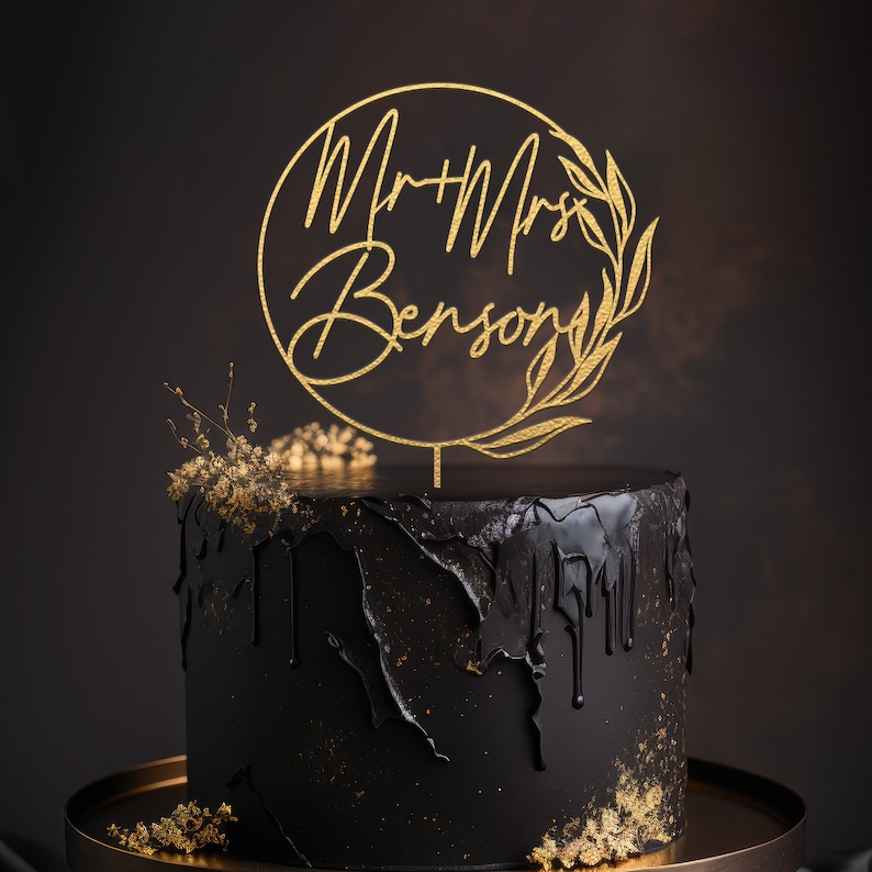 May include: A black wedding cake with gold drip icing and a gold circle cake topper with the text "Mr & Mrs Benson" and a leafy design.
