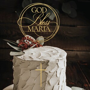 Peut inclure: Un g&acirc;teau blanc avec un gla&ccedil;age dor&eacute; et une croix dor&eacute;e sur le dessus. Un topper de g&acirc;teau dor&eacute; avec le texte "GOD Bless MARIA" est sur le dessus du g&acirc;teau.