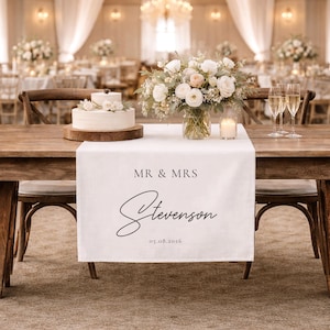 May include: A white linen table runner displays "MR & MRS Stevenson" and the date 05.08.2026. The table is set with a cake, floral arrangements, candles, and champagne flutes. The scene is set for a wedding reception.