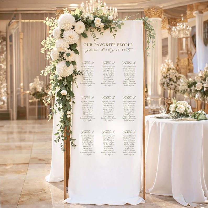 Pu&ograve; includere: Un alto cartello bianco per il piano dei posti a sedere di un matrimonio su una cornice di legno, decorato con fiori bianchi e fogliame verde. Il cartello riporta il testo "OUR FAVORITE PEOPLE" e "please find your seat" con nove tavoli.