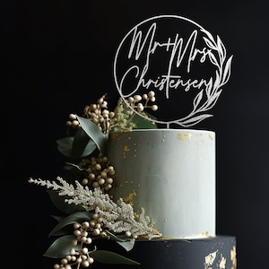 May include: A two-tiered wedding cake decorated with gold leaf and greenery. The top tier is light blue and the bottom tier is black. A silver cake topper with the text "Mr & Mrs Christensen" is on top of the cake.