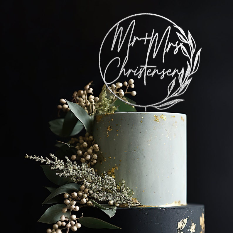 May include: A two-tiered wedding cake decorated with gold leaf and greenery. The top tier is light blue and the bottom tier is black. A silver cake topper with the text "Mr & Mrs Christensen" is on top of the cake.