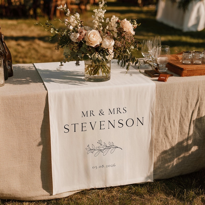 May include: A cream table runner with the text "MR & MRS STEVENSON" in black, a floral design, and the date 05.08.2026. A floral arrangement in a glass vase sits on the table, with wine glasses and a wooden serving tray.