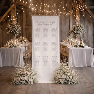 Pu&ograve; includere: Un alto cartello verticale bianco per il tableau de mariage su un supporto in metallo dorato. Il cartello presenta il testo "OUR FAVORITE PEOPLE" e "please find your seat" sopra un elenco di nove tavoli con nomi in caratteri neri.