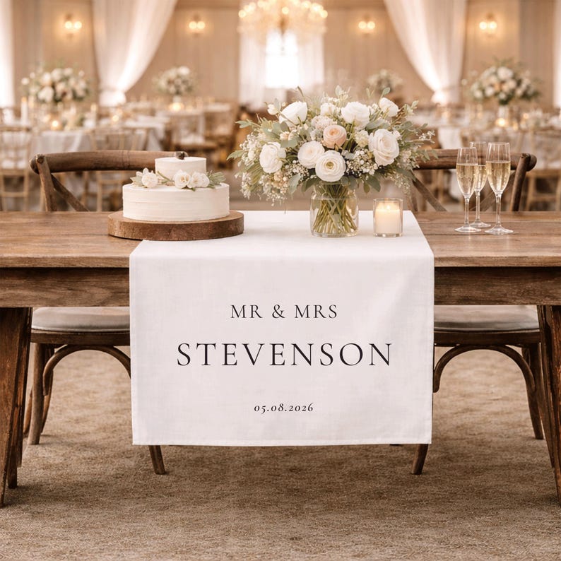 May include: A white linen table runner with the text "MR & MRS STEVENSON 05.08.2026" on a wooden table. The table is set for a wedding reception, with a cake, flowers, and champagne glasses. The room is decorated for a wedding.