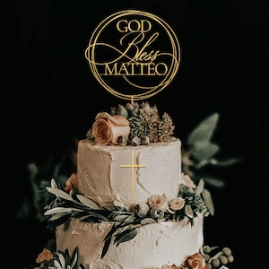 Peut inclure: Un g&acirc;teau blanc &agrave; deux &eacute;tages d&eacute;cor&eacute; de verdure et de fleurs. Un ornement dor&eacute; avec le texte "God Bless Matteo" est plac&eacute; sur le dessus du g&acirc;teau.